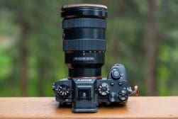 A digital camera with a zoom lens mounted on a wooden surface, set against a blurred green background.