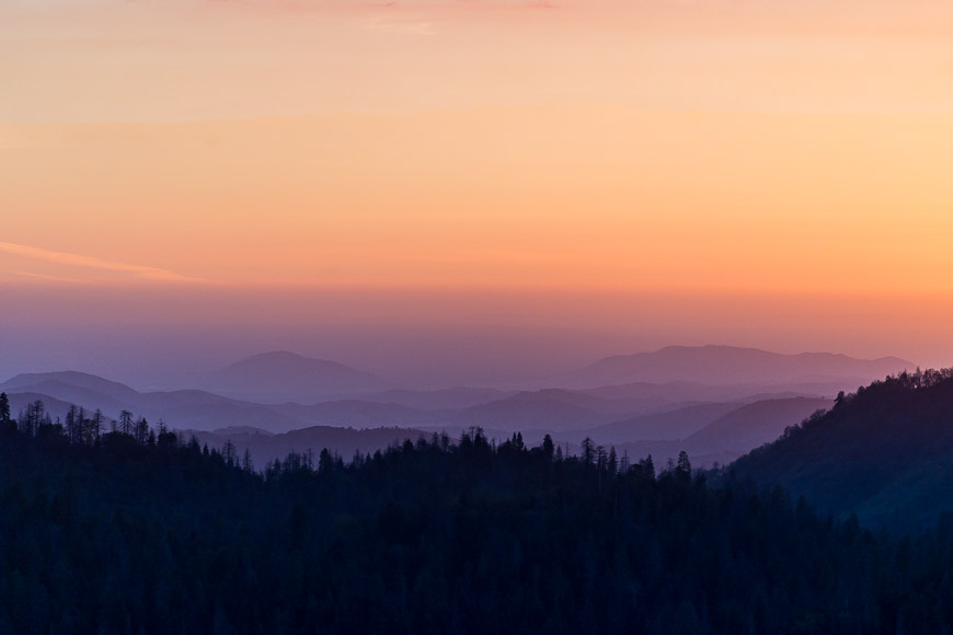 A serene mountain landscape at sunrise, featuring layers of silhouetted hills and a gradient sky transitioning from deep blue to orange.