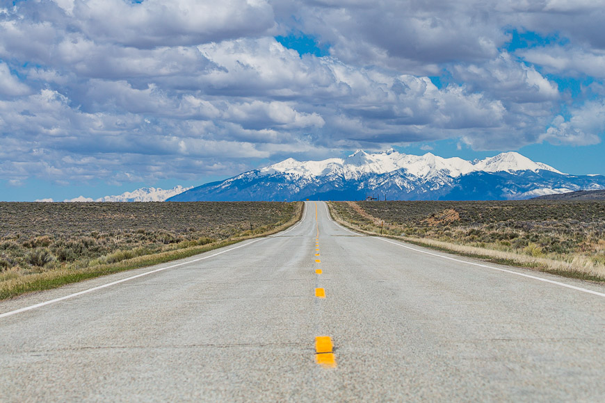 A straight road leads to snow-capped mountains under a partly cloudy sky. The road is flanked by arid open land.
