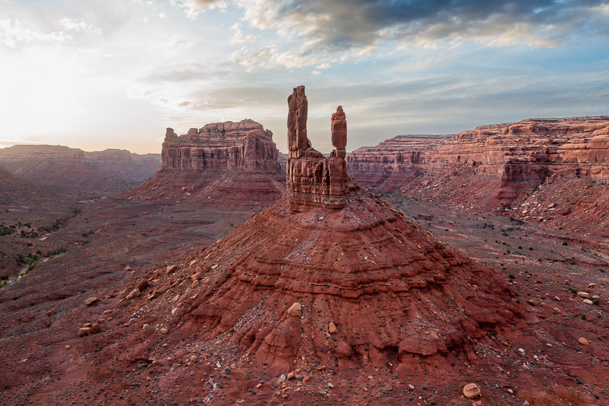 A landscape view of a desert with red rock formations, including three tall spires in the center, under a cloudy sky.