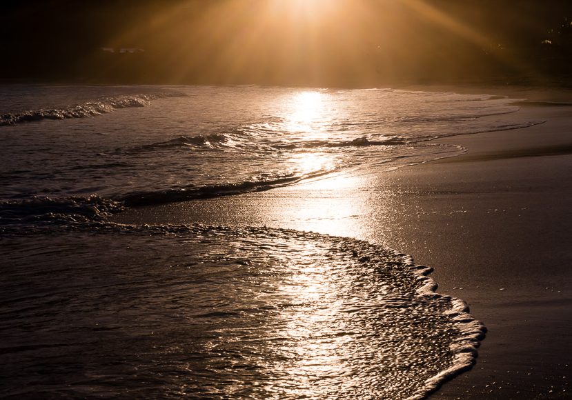 A serene beach scene with gentle waves washing onto the shore at sunset. The sun is low on the horizon, casting a golden reflection on the wet sand and water. A serene beach scene with gentle waves washing onto the shore at sunset. The sun is low on the horizon, casting a golden reflection on the wet sand and water.