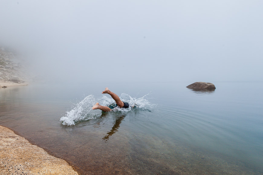 A person swimming near the shore in a foggy lake, creating splashes. A large rock is visible in the calm water.