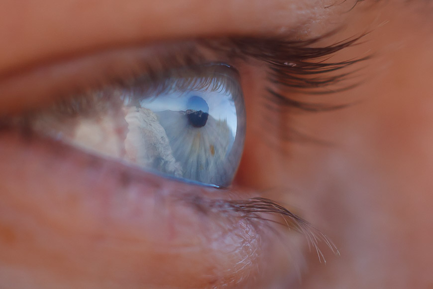 Close-up of an eye reflecting an outdoor scene, possibly a snowy mountain or glacier landscape. The image captures the vibrant blue hue of the iris and the fine details of the eyelashes.