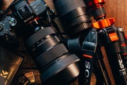 Close-up of a Sony camera with attached lens, two additional lenses, a red-handled tripod, and memory cards on a wooden surface.