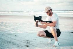 A person crouches on a sandy beach while holding a camera mounted on a stabilizer. The ocean and sky are visible in the background.