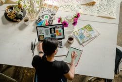 A person working at a desk with a laptop, tablet, and notebooks, surrounded by art supplies, flowers in a vase, and a cup of coffee.